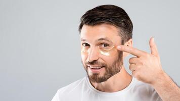 Smiling handsome man applying eye patches on his face, closeup. Middle-aged bearded man with golden patches under eyes from wrinkles and dark circles, grey studio background, face care for men concept photo