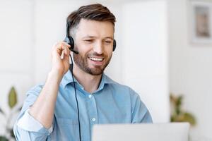 Hotline Operator. Handsome Young Man Customer Support Manager In Headset Working With Laptop Computer In Office, Consulting Clients Online, Managing Issues, Sitting At Desk, Free Space photo