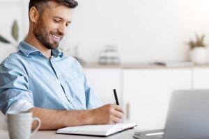 Smiling Handsome Businessman Taking Notes To Notepad At Workplace In White Office, Noting Business Ideas, Planning Budget And Working Schedule, Sitting At Desk With Laptop Computer, Free Space photo
