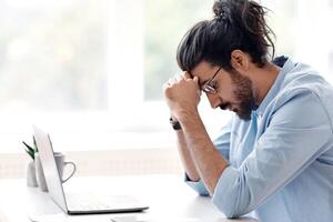 Upset Eastern Freelancer Guy Suffering Lack Of Inspiration While Working On Online Project With Laptop At Home, Sitting At Desk Near Window And Touching Head In Despair, Side View With Copy Space photo