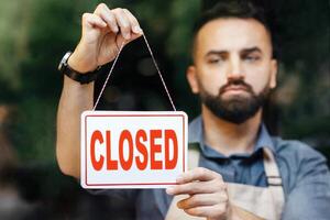 Closure of small businesses, problems in flowers shop during covid-19 quarantine. Sorrowful millennial bearded caucasian man in apron turns nameplate is closed, in store with different plants photo