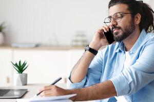 Busy Western Male Entreprener Talking On Mobile Phone And Taking Notes While Working In Office, Writing In Notepad, Sitting At Desk And Talking With Client, Planning Business Schedule, Free Space photo