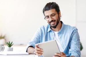 Arab Freelancer Guy Using Digital Tablet At Home, Having Break In Work, Sitting At Desk, Browsing Internet Or Social Media, Checking New App, Smiling Eastern Man Messaging Or Shopping Online photo