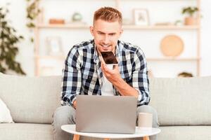 Voice Search. Portrait of guy talking on smartphone or using virtual personal assistant app on his device sitting on the couch in living room, working on laptop. Man recording audio message or command photo
