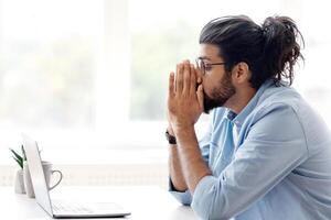 Stressed Arab Freelancer Guy Suffering Lack Of Ideas While Working On Laptop At Home, Sitting Upset At Desk With Computer, Depressed Eastern Man Holding Hands Near Face, Side View With Copy Space photo