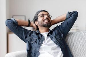 Relaxed smiling arab guy leaning back on couch at home, relaxing after successful day, positive young eastern man resting on sofa with hands behind head, enjoying domestic weekend, free space photo