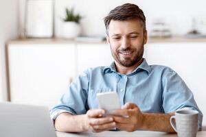 Happy Millennial Businessman Relaxing With Smartphone At Workplace In Office, Leaning Back In Chair, Texting With Friends Or Browsing Internet, Having Break In Work, Sitting At Desk With Laptop photo