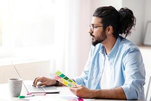 Young Eastern Freelance Graphic Designer Guy Working With Laptop And Color Swatches, Sitting At Desk In Home Office, Choosing Colour Gamma For New Design Project, Side View With Copy Space photo