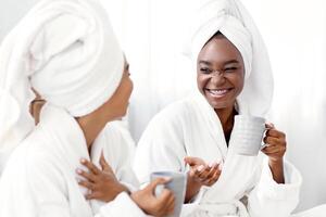 Two joyful african american girlfriends in bathrobes joking and drinking coffee. Cheerful black women with towels on their head having spa day at home, drinking tea and having funny conversation photo