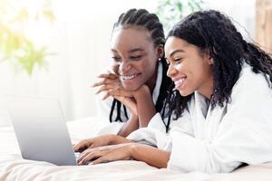 Two happy black women laying on bed and using laptop at home, websurfing, empty space. Smiling african american girlfriends enjoying weekend, checking social media together on computer photo