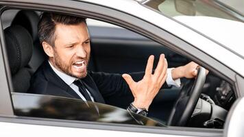 Expressive middle-aged businessman screaming and gesturing while driving car, side view, through window shot. Furious entrepreneur stuck in traffic, getting late for business meeting photo