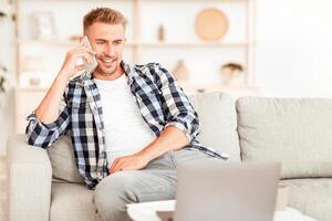 Phone Call. Portrait of confident adult man talking on cellphone, sitting on the couch in living room, looking at computer. Communication, Rest And Relax Concept. Copy space. Guy having conversation photo