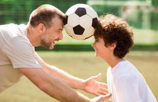 Side view portrait of mature father with his little son playing soccer on football pitch, holding ball with head photo