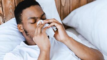 Ill Black Man Blowing Runny Nose In Paper Tissue Having Rhinitis Lying In Bed At Home. Sick African Guy Sneezing Having Sinusitis, Flu And Cold Symptom Concept. Panorama photo