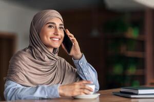 sonriente bonito musulmán dama hablando en móvil teléfono mientras descansando a café solo, gratis espacio. joven contento árabe mujer en hijab teniendo conversacion en teléfono inteligente y Bebiendo té o café foto