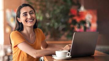 Young smiling arab woman using laptop while sitting at cafe alone, empty space. Female freelancer working online, typing on keyboard, sending email to client or chatting, drinking coffee or tea photo