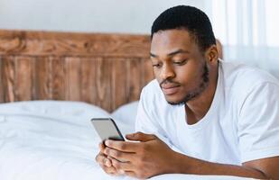Serious Black Man Using Mobile Phone Browsing Internet Reading Online News On Smartphone Lying In Bed At Home. Cellphone Application Concept. Empty Space For Text photo