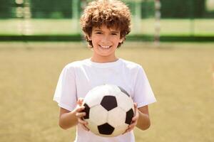 Portrait of cute curly boy holding soccer ball on the football field and looking at camera, posing, selective focus photo