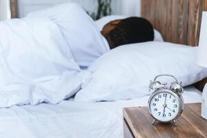 Oversleeping. Overslept Black Man Sleeping Through Alarm Clock In The Morning Lying In Bed At Home, Focus On Alarm-Clock. Tardiness, Punctuality, Early Awakening Concept. Shallow Depth photo