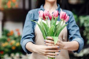 Spring bloom, creating bouquets for holiday and congratulations in plants shop. Happy young beautiful woman in apron holds tulips for client, on flower studio background, cropped, copy space photo