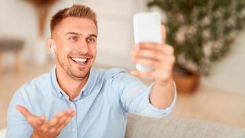 Online Call. Portrait of smiling man making virtual conference, holding mobile phone, wearing wireless earbuds, blurred background. Communication Concept. Guy chatting using smartphone photo