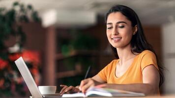 Working female student spending time at cafe, taking notes at notepad, using laptop and drinking coffee, panorama with copy space. Smiling young lady working online at cafeteria, side view photo