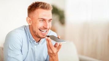 Voice Search Concept. Closeup portrait of young bearded guy using voice assistant or calling on smartphone at home, blurred background, copy space. Smiling man talking on mobile phone photo