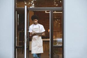 New normal and opening of cafe after covid-19 quarantine. Millennial attractive african american man in apron working on digital tablet near front door of modern cafeteria, empty space photo