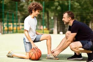 Preparation. Portrait of supportive man tying shoelaces of curly boy on basketball playground, blurred background photo