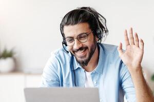 Distant Communication. Happy Western Man In Headset Waving Hand At Laptop Camera While Making Call Via Computer In Office, Chatting With Family Or Having Online Conference, Copy Space photo