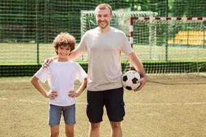 Sports Concept. Portrait of smiling man holding soccer ball and standing with boy on playground, posing at camera photo
