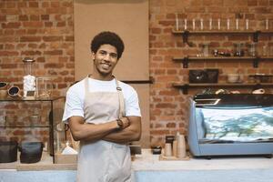 Modern coffee shop open after lockdown. Smiling young african american guy in apron, with crossed hands near bar counter with coffee equipment and looks at camera, waiting for clients, free space photo