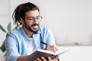 Smiling Western Freelancer Guy Taking Notes To Notepad At Workplace In Home Office, Planning Working Schedule, Filling Diary, Noting Business Ideas, Sitting At Desk With Laptop Computer, Copy Space photo