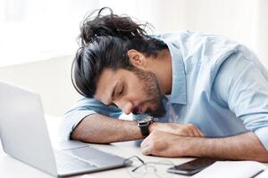 Sleeping At Work. Overworked freelancer guy napping at workplace at home office, tired after working on online project with laptop, young eastern man taking a nap at desk, placed head on hands photo
