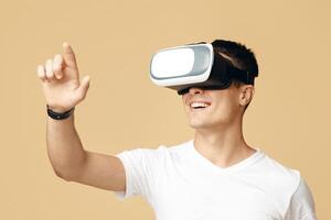 Man have fun with virtual reality headset. Cheerful young guy student in white t-shirt and virtual reality glasses touches virtual button on empty space isolated on sand background, studio shot photo