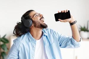 Benefits Of Freelancing. Cheerful Arab Guy Listening Music And Singing While Working At Home Office, Joyful Man Using Cellphone As Microphone, Enjoying Self-Employment And Remote Job, Free Space photo