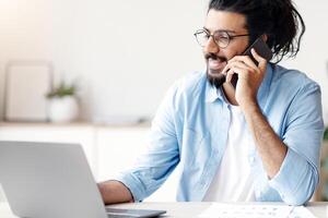 Business Communication. Young Indian Male Entrepreneur Talking On Cellphone While Working On Laptop At White Office, Looking At Computer Screen And Smiling, Discussing Project By Phone, Copy Space photo