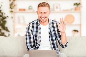 Online Call. Portrait of smiling guy making virtual conference on laptop, waving with hand to webcam. Communication Concept. Man chatting using computer, sitting on the couch at home photo