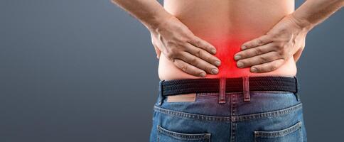 Closeup of unrecognizable man in jeans rubbing his low back, having pain, sore zone highlighted with red, grey studiio background. Backache, rheumatism, osteochondrosis, radiculitis concept photo