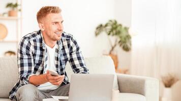 Home Office. Happy casual man holding smartphone and using laptop, daydreaming and looking away at copy space. Successful professional guy working remotely and thinking about business and future photo