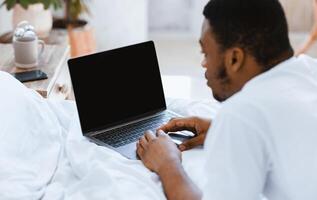 Unrecognizable African American Freelancer Man Using Laptop Computer With Blank Screen, Working Online And Advertising Internet Website Lying In Bed At Home. Mockup, Selective Focus photo