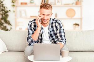 Communication Concept. Smiling millennial man talking on the cellphone using laptop at home office, happy young guy making phone call having mobile conversation looking at computer sitting on sofa photo