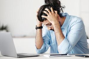 Work Problems. Desperate indian manager sitting at desk in office, suffering from business failure, having lack of inspiration or deadline stress, depressed western man touching head, free space photo