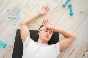 Fatigue after exercising at home. Tired young man wipes sweat, lying on mat on floor with sports tools and empty bottle for water, top view, free space photo