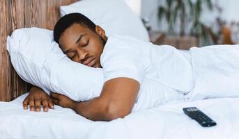 Fast Asleep Black Man Sleeping Embracing Pillow Resting In Bed At Home, Lying Near Remote Control Tired After Bingewatching TV Shows At Night. Rest, Napping And Recreation Concept. photo