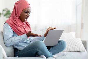 Computer Problems. Angry black muslim lady in hijab having troubles with laptop at home, annoyed african woman looking at device screen and emotionally gesturing, having bad internet connection photo