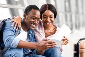 Waiting For Flight. Happy Black Couple Using Smartphone In Airport , Having Fun In Terminal Together Before Flight Boarding, Holding Passports Tickets And Taking Selfie With Mobile Phone Camera photo