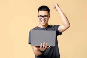 Great job, solving problem and win in game. Happy young guy student with glasses looks at laptop and expresses emotions of victory, isolated on beige background, studio shot photo