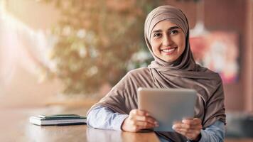 Happy girl in hijab with digital tablet smiling at camera, sitting at cafe alone, copy space photo