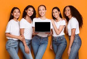 Five Multicultural Women Showing Laptop Empty Screen Recommending Website Standing Over Yellow Studio Background, Smiling To Camera. Females Group Showing Computer Display Posing Together. Mockup photo